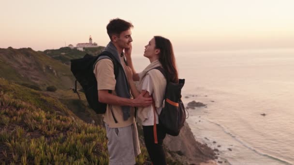 一对背着背包的年轻夫妇在悬崖边接吻 日落时俯瞰大海 标志着一次风景远足的结束 — 图库视频影像
