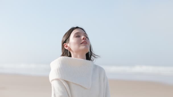 在一个阳光明媚而寒冷的海滨日子里 一个穿着白色毛衣 紧闭双眼 享受着清爽的海风的年轻女子的画像 — 图库视频影像