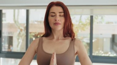 Huzurlu meditasyonda kızıl saçlı yetişkin kadın namaste 'ye poz veriyor, gözleri kapalı, gözleri açık, sakin bir stüdyoda sükunet içinde..