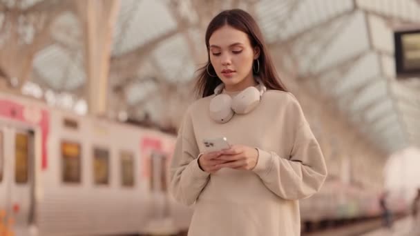 集中精神的年轻女性 带着耳机 在火车站用手机 可能会在出发前搜寻她的站台或行程 — 图库视频影像