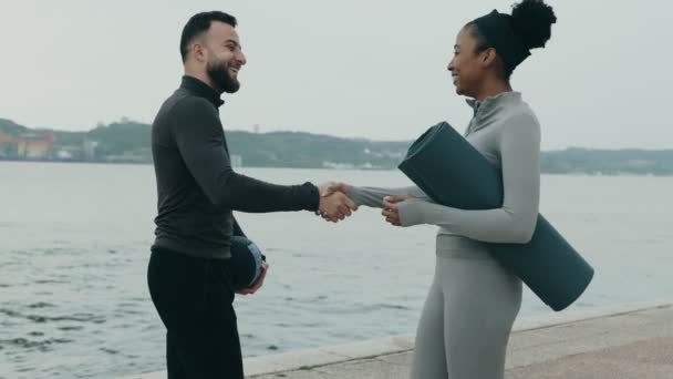 男性のフィットネストレーナーは 薬ボールを持つ女性のクライアントにヨガマットを開催し 屋外のパーソナルトレーニングセッションの前に手を振ります — ストック動画