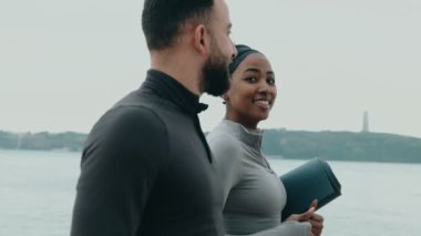 Genç siyahi kadın ve beyaz adam yan yana yürüyorlar, yoga minderi tutuyorlar, su kenarında spor salonuna başlamaya hazırlar..