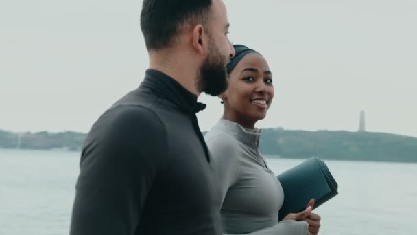 フィット若い黒人女性と慎重な男性は横に歩き ヨガマットを保持し 水で屋外フィットネスセッションを開始する準備ができています — ストック動画