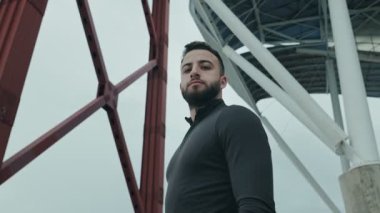 Spor giyimli kararlı genç adam bir köprünün altında güven içinde duruyor ve egzersiz rejimine hazırlanıyor. Düşük açıyla yakalanmış, güç ve motivasyon çağrıştırıyor..