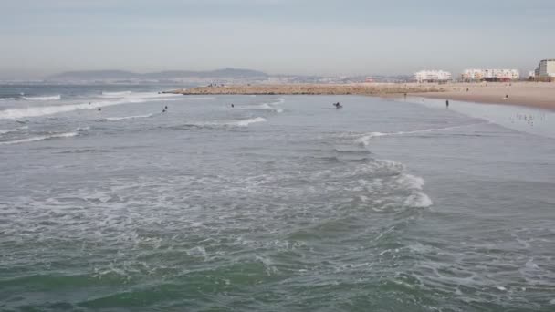 在平静的海滩上 冲浪者乘着柔和的海浪 背景是清澈的天空下的海滨城市景观 传达着一种宁静和休闲的感觉 — 图库视频影像