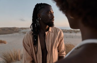 Afro örgü saç stili ve yaz elbisesi giymiş genç Afrikalı Amerikalı erkek, kumlu tarlada kuru otlarla dikilirken yüzünü çeviriyor. Bulutlu mavi gökyüzüne karşı isimsiz bir kadın.