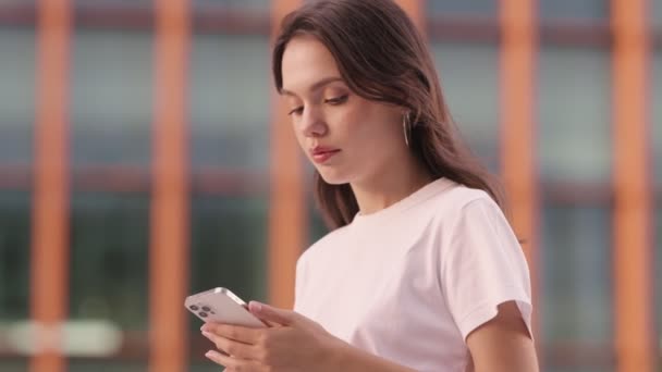 一个年轻的女人专注于她的智能手机 可能在等待一个消息或一个朋友 在一个模糊的城市背景下设置场景 — 图库视频影像