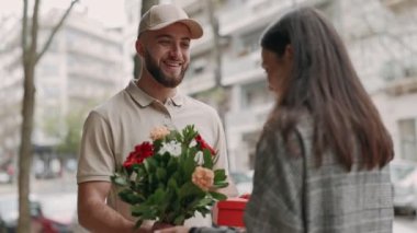 Şapkalı gülümseyen teslimatçı güzel bir çiçek buketi ve şehir sokaklarında mutlu bir genç kadına kırmızı bir hediye kutusu veriyor..