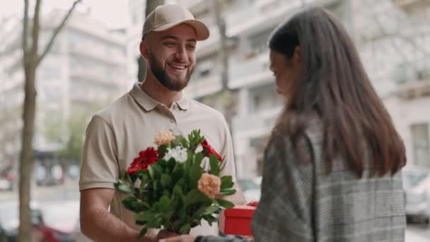 美しい花束と赤いギフトボックスを提供するキャップの笑顔の配達男性 都市の通りで幸せな若い女性に — ストック動画