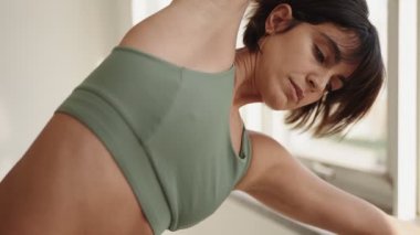 Yeşil aktif giysili, odaklanmış esmer bir kadın pencerenin kenarına doğru incelikle uzanır. Yoga rutinine evde sakin bir şekilde hazırlanır..