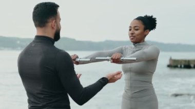 Beyazlı bir erkek eğitmenle açık havada yapılan bir eğitim, genç bir siyahi kadına halter egzersizlerinde rehberlik ediyor.