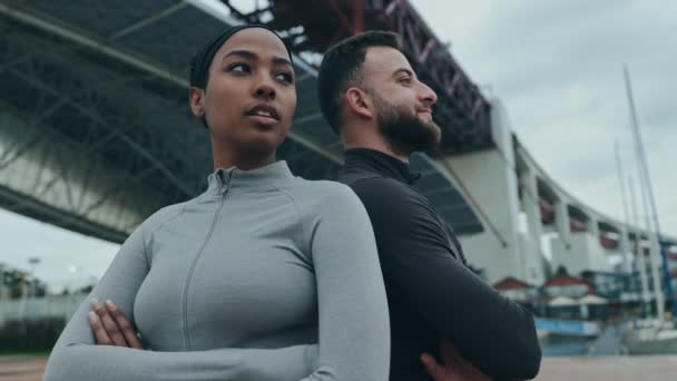 一名健康的黑人女子和一名身穿运动服的肌肉发达的高加索男子背靠背地站着 表情坚定 在室外展示出多样性和团队精神 — 图库视频影像