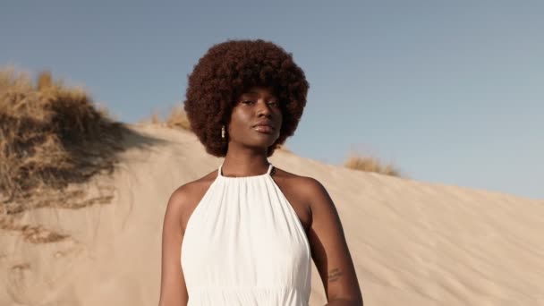 エレガントなアフリカの女性は自信と自然の美しさを誇り 砂丘の背景にシックな白いドレスを飾り スタイルと洗練の本質を捉えています — ストック動画