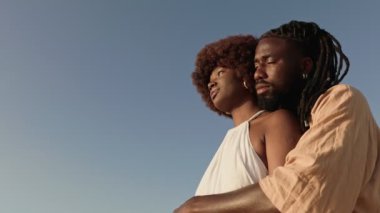 Romantik Afrikalı çift açık havada sevgiyle kucaklaşıyorlar. Arka planda açık mavi bir gökyüzü var. Hem moda hem de şefkat gösteriyorlar..