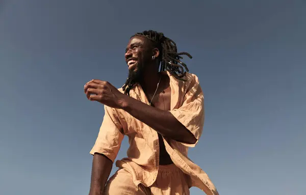年轻的非洲裔美国男子面带微笑 头戴非洲式辫子 穿着夏装 在阳光灿烂的阳光下 独自站在无云的蓝天下 望着远方 — 图库照片