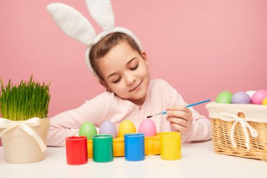 Küçük kız masada oturan, pembe arka plan Paskalya kutlamaları için yumurta boyama içerik