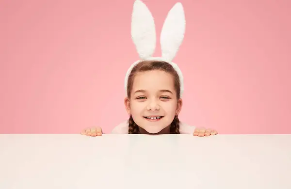 可爱的女孩穿着复活节兔子的白色耳朵 而坐在白色的桌子后面仔细地看了出来 — 图库照片