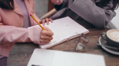 İşbirlikçi bir proje planlama oturumu sırasında, arkadaşlarıyla birlikte, bir günlüğe notlar yazan genç bir kadının elini çeken odaklanmış bir çekim..