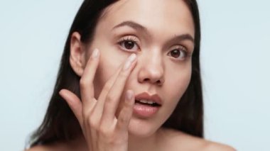 Doğal makyajlı genç bir kadının, pürüzsüz cildine dokunuşunun yakın plan resmi. Düz mavi arka plan, cilt bakımını ve güzelliğini sembolize ediyor..