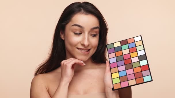 カラーメイクパレットを持っている笑顔の若い女性 ベージュの背景で創造性と選択を表現 — ストック動画