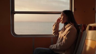 Bir feribotun penceresinde oturan genç kadın, güneş batarken altın rengini yansıtıyor..
