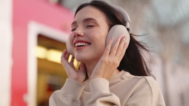 一位快乐的年轻女子 紧闭双眼 一边听着耳机里的音乐 一边感受着音乐的节奏 一边在室外表达着快乐与满足 — 图库视频影像