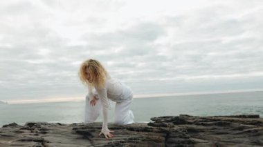 Beyazlar içindeki bir kadın kayalık sahilde özgürce dans ediyor. Uçsuz bucaksız okyanusun yanında huzur ve özgürlük hissi uyandırıyor..