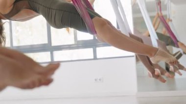 Bir grup yetişkin kadın, parlak bir stüdyoda mor ve beyaz hamakları kullanarak havadan yoga dersinden zevk alıyorlar..