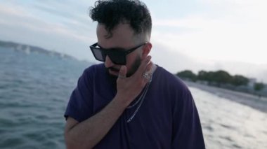 Güneş gözlüklü, düşünceli, sakallı bir adam, sakin bir ifadeyle denizin kenarında duruyor, yüzüne nazikçe dokunuyor, sakinlik hissi uyandırıyor..