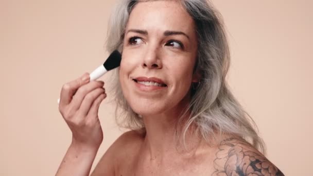 タトゥーと灰色の髪を持つ50代の優雅な女性は ベージュの背景に隔離されたメイクブラシで基礎を適用します — ストック動画
