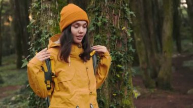Sarı ceketli ve turuncu bereli bir kadın yoğun, yeşil bir ormanı keşfetmeden önce sırt çantasını düzeltiyor..