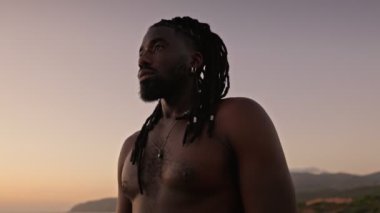 Üstsüz Afro-Amerikalı bir adam, rastalı saçlarıyla gün batımına karşı duruyor, doğal ortamda dinginlik ve güç yayıyor..