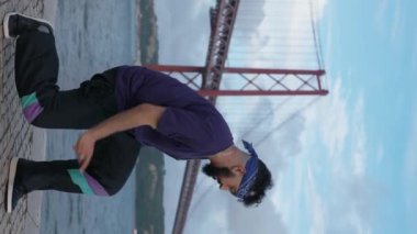 Moda sokak giyiminde engelli neşeli ve iri bir adamın dikey görüntüsü dans yoluyla neşe ve özgürlüğü ifade ediyor, arka planda manzaralı bir köprü var..