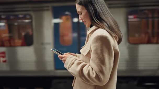 地下鉄駅で地下鉄を待っている間に彼女の携帯電話に従事したベージュコートの若い女性の側面 — ストック動画
