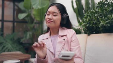 Kafedeki Asyalı genç bir kadın akıllı telefonuyla şarkı seçiyor, bitkilerin arasında gürültü önleyici kulaklıklarıyla müzik dinliyor..