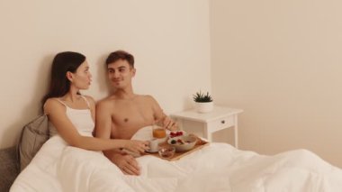 Sevecen bir çift yatakta bir kahvaltı tepsisiyle romantik bir anı paylaşıyor. Sıcak bir yatak odasında sevgi, yakınlık ve rahatlık yayıyorlar..