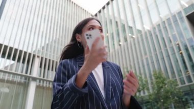 Takım elbiseli profesyonel genç bir kadın, arka planda modern ofis binalarıyla telefon görüşmesi yapıyor..