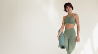 Modern aktivite kıyafetleri içinde formda genç bir kadın yoga seansına hazırlanıyor. Elinde yoga minderi var..