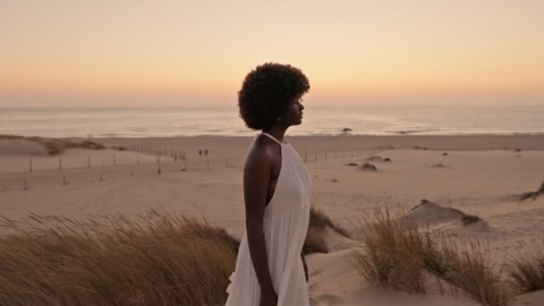 Elegancka Czarna Kobieta Płynącej Białej Sukience Stoi Plaży Zmierzchu Łagodnym — Wideo stockowe