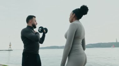Sportif bir adam deniz kenarındaki bir kadına spor kıyafetleriyle omuz eğitimi veriyor..