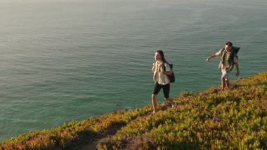 Sırt çantalı genç bir çift, sakin bir günbatımında arka planda engin bir okyanusla kıyı şeridi boyunca manzaralı bir yürüyüş yapıyor..