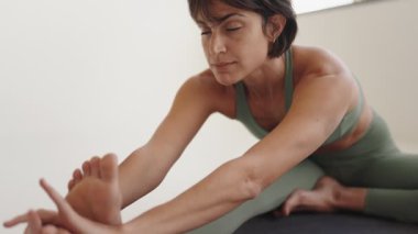 Odaklanmış genç bir kadın, güneşli, sakin bir stüdyo ortamında rahatlatıcı bir yoga esneme rutinine girişiyor..