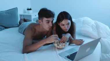 Bir çift, dizüstü bilgisayarda film izlerken yatakta dinleniyor ve geceleri patlamış mısırın tadını çıkarıyor..