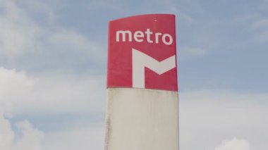 Arka planda açık mavi gökyüzü olan bir direkte canlı kırmızı metro istasyonu tabelası, şehir içi ulaşımı simgeliyor..