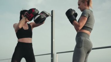 İki kararlı kadın eldiven ve pedlerle boks antrenmanına girişiyor. Açık bir zemine karşı dayanıklılık ve takım çalışması sergiliyorlar..