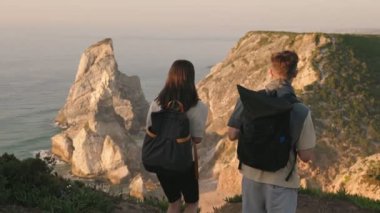 Gün batımında görkemli deniz kayalıklarına bakan, manzaralı bir yürüyüş yoluna ulaştıktan sonra zafer kazanmış bir çiftin arka görüntüsü..
