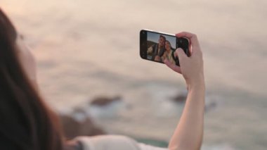 Omuz hizasında, elinde bir telefonla mutlu bir çiftin gün batımında okyanus zemininde selfie çekerken görüntüsü..