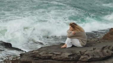 Yansıtıcı bir kadın denizin kayalık kenarında dizlerini kucaklayarak yalnız oturur güçlü dalgalara karşı düşüncelerinde kaybolur..