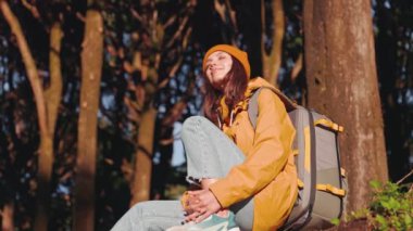 Sarı ceketli ve bereli mutlu genç bir kadın sırt çantasıyla dinleniyor. Orman ortamında güneş ışığının tadını çıkarıyor..