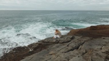 Yalnız bir kadın kayalık sahilde düşünceli bir şekilde oturur, sonsuz okyanusa bakar, sükuneti ve güzelliğin içindeki yansımayı somutlaştırır..
